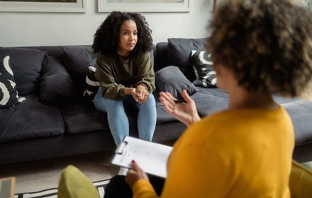 duas mulheres sentadas uma de frente para outra, uma com a prancheta na mão e outra com as mãos dadas olhando séria